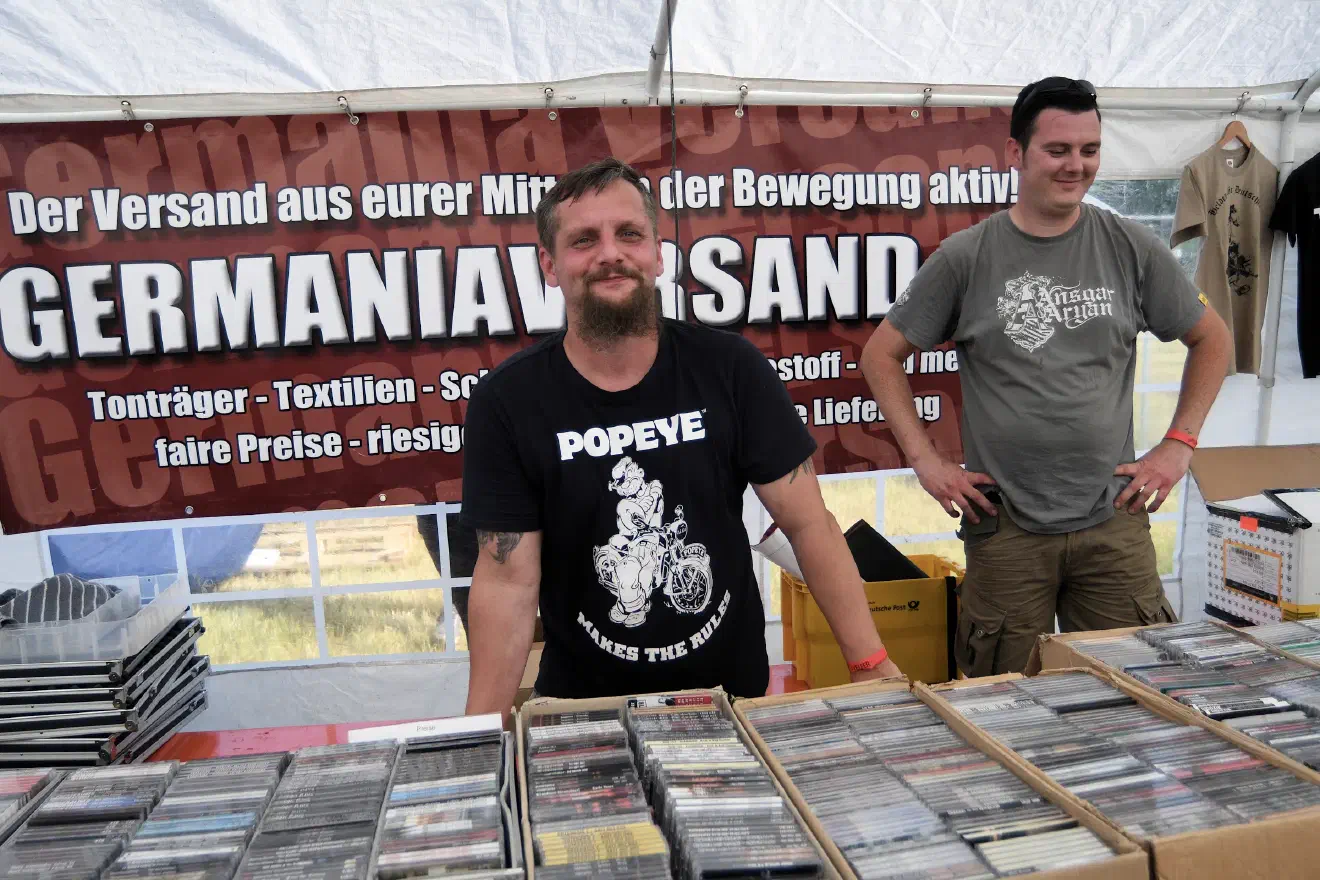Patrick Weber (hier rechts im Bild auf einem Rechtsrockfestival 2018 in Themar) verantwortet den "Germania-Versand" - einem Versandhandel, der ein neonazistisches Publikum als vordergründige Zielgruppe hat. Weber präsentiert sich dort als "Bewegungs-Unternehmer", einen Neonazi, der erwirtschaftete Mittel wieder in den Strukturaufbau fliessen lässt.