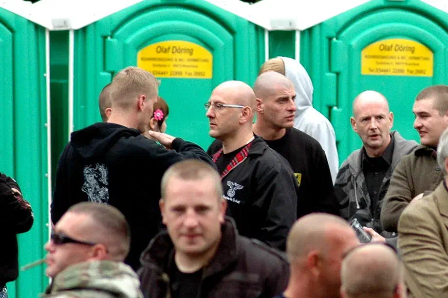 Aber auch in der Bundesrepublik kam es immer wieder zu Zusammenkünften von terroristisch agierenden Blood & Honour Aktivist*innen. So unter anderem beim sogenannten "Fest der Völker", einem Neonazifestival in Jena. Im Jahr 2007 trafen sich dort Aktivisten von Blood & Honour und Combat 18 aus verschiedenen europäischen Ländern. Unter diesen befand sich auch Illes Zsolt, Anführer von Blood & Honour Ungarn. Rechts von Zsolt findet sich ein Aktivist aus dem Umfeld der belgischen Terrororganisation "Bloed, Bodem, Eer & Trouw" (BBET)