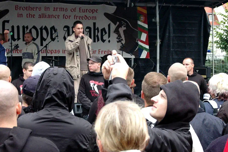 Ein weiter Neonazi, der sich derzeit als Unterstützer des "Nationalsozialistischen Untergrunds" (NSU) vor dem Oberlandesgericht München wegen Beihilfe zum Mord verantworten muss, war damals in Jena zugegen. Rald Wohlleben (Bild: auf der Bühne) steht im Verdacht Rechtsrock-Konzerte für die Terrororganisation NSU organisiert zu haben. Mit den erzielten Einnahmen sollte eine Flucht nach Südafrika finanziert werden. Wohlleben wird außerdem vorgeworfen, Beihilfe zu sechs Morden geleistet zu haben, indem er "dem NSU 2001 oder 2002 eine Schusswaffe nebst Munition" verschaffte, die ein Kurier "in seinem Auftrag zu den NSU-Mitgliedern brachte". "Aufgrund seiner anhaltenden Verbindung zu der unter falscher Identität lebenden Gruppe" soll er von den terroristischen Straftaten gewusst haben, so die Bundesanwaltschaft.